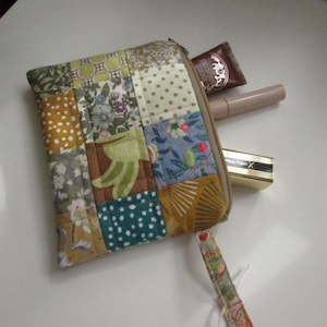 May include: A patchwork cosmetic bag with a zip, made from squares of patterned fabric in green, yellow, blue, and brown. The bag is open, showing a lipstick, a mascara tube, and a small brown packet.