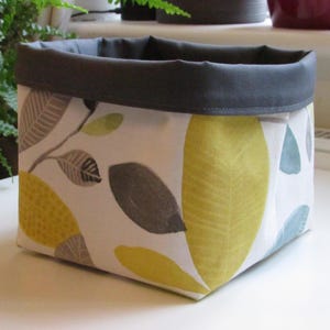 May include: A fabric storage basket with a white background and a leaf pattern in yellow, grey, and blue. The top edge is a solid dark grey. The basket is on a white surface.