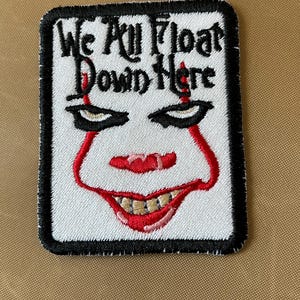 May include: White embroidered patch with a black border. The patch features a red and white clown face with the text "We All Float Down Here" in black.