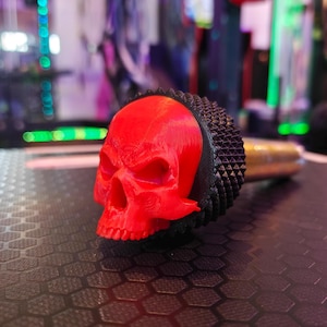May include: A red skull-shaped gear shift knob with a black, textured base. The knob is made of 3D printed plastic.