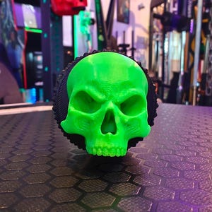 May include: A green skull-shaped object with a black textured base. The skull has a menacing expression with deep-set eyes and a prominent jawline.