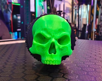 May include: A green skull-shaped object with a black textured base. The skull has a menacing expression with deep-set eyes and a prominent jawline.