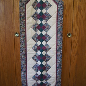 May include: A quilted table runner with a cream-colored center and a diamond pattern in teal, purple, and cream. The edges are bordered with a patterned fabric in similar colors. The runner is hanging on a wooden door.