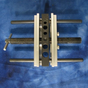 May include: A metal tool with a black and silver design. The tool has multiple holes and rods, with a textured handle and a screw mechanism. The tool is designed for mechanical work.