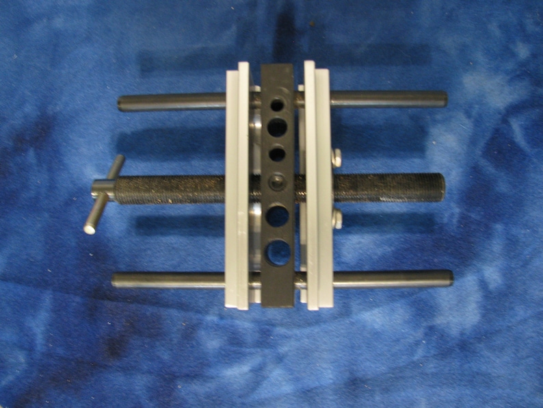 May include: A metal tool with a black and silver design. The tool has multiple holes and rods, with a textured handle and a screw mechanism. The tool is designed for mechanical work.