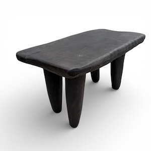 May include: A dark brown, rectangular wooden stool with three rounded legs. The top surface has a slightly uneven texture and a natural, rustic appearance. The stool is a solid, handcrafted piece.