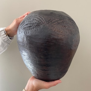May include: A large, dark brown ceramic vase with a rounded, bulbous shape. The vase has a textured surface with carved lines and patterns.