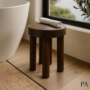 May include: A dark brown wooden stool with four legs and a round top. A white towel is rolled up and placed on top of the stool.
