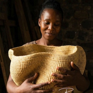 May include: A large, hand-woven basket made from natural light-tan fibers, featuring a wide, undulating, and organic rim shape with a dense, uniform weave pattern throughout the body of the container.