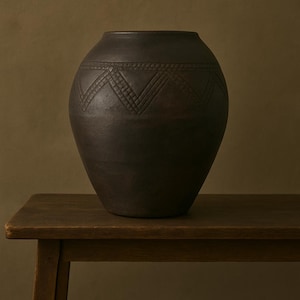 May include: A dark brown ceramic vase with a geometric pattern around the middle. The vase is sitting on a wooden table.
