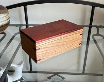 May include: A rectangular wooden box with a reddish-brown lid and a light-colored body. The box is made of layered wood and has dark accents at the corners. It sits on a glass surface.