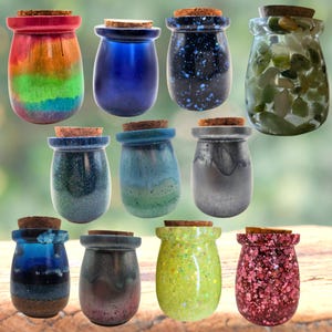 Puede incluir: Una colección de once frascos pequeños y redondeados con tapones de corcho, que presentan varios acabados, incluidos degradados de arcoíris, azul sólido, azul oscuro con purpurina, vidrio transparente lleno de piedras verdes, purpurina azul verdoso, degradado azul-verde, gris y purpurina rosa.