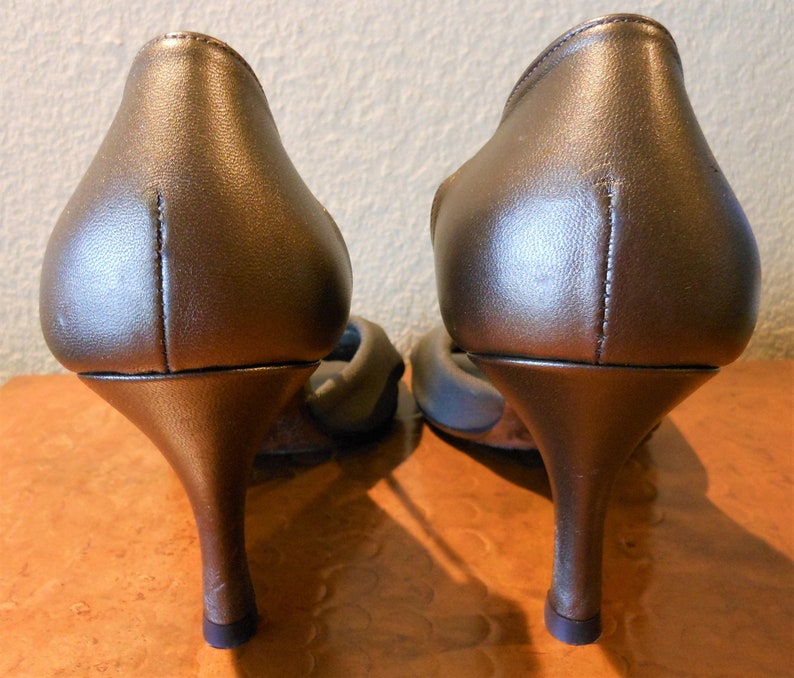 May include: A pair of bronze-colored high-heeled shoes. The shoes have a pointed toe and a thin, curved heel. The heels are approximately 3 inches tall.