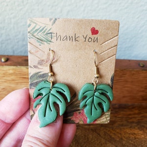 May include: A pair of green monstera leaf dangle earrings with gold hooks. The earrings are displayed on a brown card with the text "Thank you" and a red heart.
