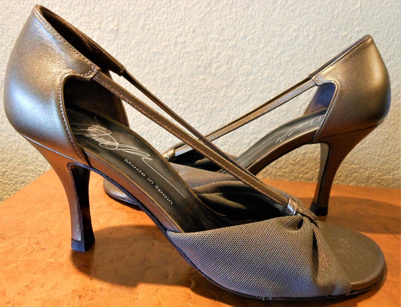 May include: A pair of bronze-colored high-heeled shoes with a slingback design. The shoes have a pointed toe and a thin strap that crosses the foot. The heels are approximately 3 inches tall. The shoes are made in Spain.