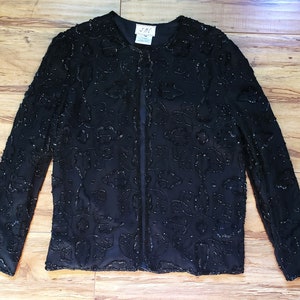 May include: A black, long-sleeved jacket with a floral beaded design. The jacket has a round collar and a straight, open front. The beading covers the entire jacket, creating a textured appearance. The jacket is displayed on a wooden surface.