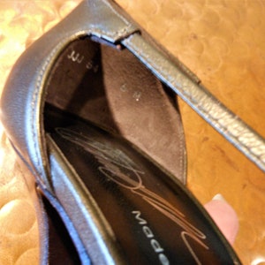May include: Close-up of a metallic silver high-heeled shoe. The shoe has a strap and a pointed toe. The interior of the shoe is dark brown, with the size marked as 6 M. The text "JJJ 54" is visible.