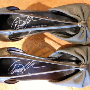 May include: A pair of metallic gold-colored flats with a bow detail on the vamp. The shoes have a slingback design and a signature on the insole. The words "Made in Spain" are printed on the insole.