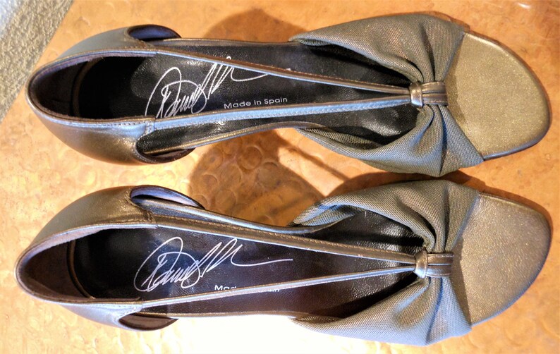 May include: A pair of metallic gold-colored flats with a bow detail on the vamp. The shoes have a slingback design and a signature on the insole. The words "Made in Spain" are printed on the insole.