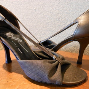 May include: A pair of bronze-colored high-heeled sandals. The sandals feature a bow detail on the toe and thin straps. The words "Made in Spain" are printed on the insole.