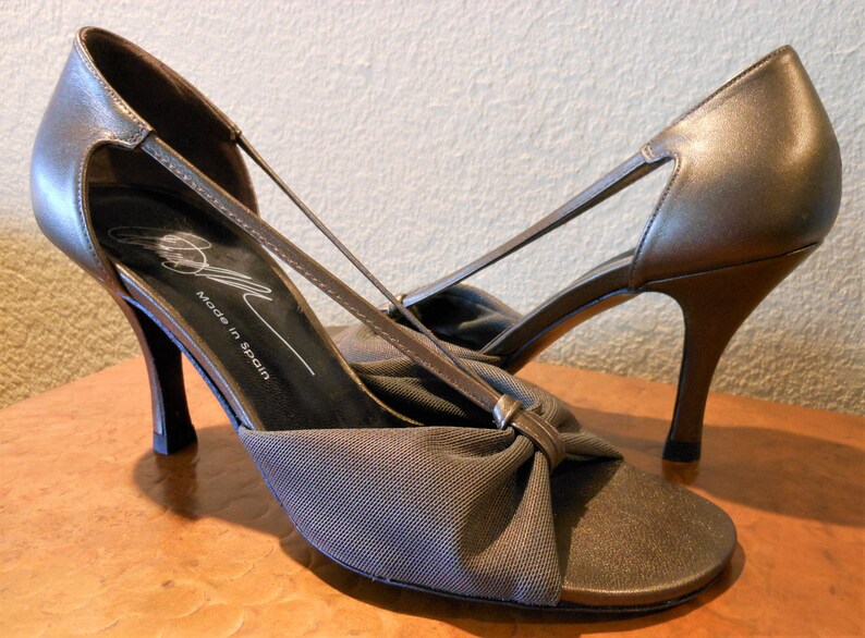 May include: A pair of bronze-colored high-heeled sandals. The sandals feature a bow detail on the toe and thin straps. The words "Made in Spain" are printed on the insole.