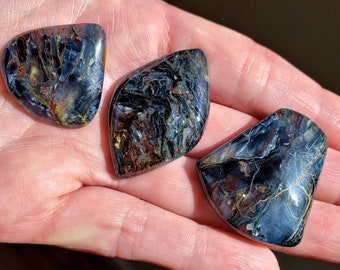 May include: Three polished pietersite gemstones with swirling patterns of blue, brown, and gold. The stones have irregular shapes and are displayed in a hand, showcasing their natural beauty and unique color variations.