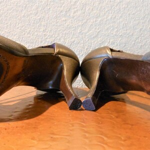 May include: A pair of bronze-colored high-heeled shoes with open toes. The soles are dark brown with a stitched border and a signature. The heels are curved and slender. The shoes are displayed on a light brown surface.