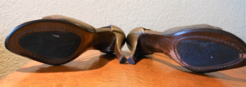 May include: A pair of bronze-colored high-heeled shoes with open toes. The soles are dark brown with a stitched border and a signature. The heels are curved and slender. The shoes are displayed on a light brown surface.