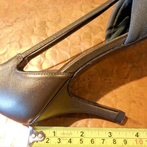 May include: Close-up of a bronze-colored high-heeled shoe with a strap and a thin heel. The shoe measures approximately 3 inches in length. The shoe is on a brown surface.