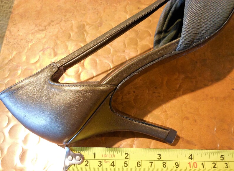 May include: Close-up of a bronze-colored high-heeled shoe with a strap and a thin heel. The shoe measures approximately 3 inches in length. The shoe is on a brown surface.