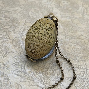 May include: A brass oval locket necklace with a floral and bird design. The locket opens to reveal a small compartment inside.