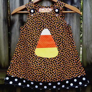 May include: A sleeveless Halloween dress with a candy corn design. The dress features a black background with candy corn print, a large candy corn applique, and a black polka dot hemline. It hangs on a wooden hanger.