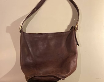 May include: A dark brown leather bucket bag featuring a smooth, rounded base and a single adjustable shoulder strap. The bag is secured with gold-toned metal hardware, including buckles on the sides that connect the strap to the main body.