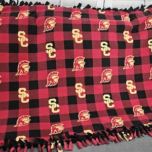 May include: Red and black plaid fleece blanket with a repeating pattern of a Trojan helmet and the letters "SC" in gold. The blanket has a fringed edge.