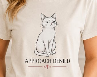 May include: A cream-colored t-shirt featuring a graphic illustration of a white cat with a stern facial expression. Below the cat, the text "APPROACH DENIED" is printed in black capital letters, accompanied by a small decorative line element.