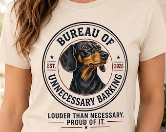 May include: A cream-colored t-shirt featuring a circular graphic with a black and tan dachshund. The text reads BUREAU OF UNNECESSARY BARKING, EST. 2026, and LOUDER THAN NECESSARY. PROUD OF IT. The design uses navy blue and red accents with star details.