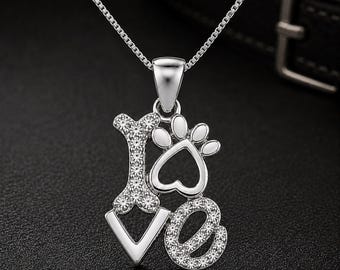 May include: A silver-toned pendant necklace featuring the word LOVE, where the letter L is shaped like a dog bone, the letter O is a paw print with a heart-shaped center, and the letters V and E are encrusted with small clear sparkling stones.