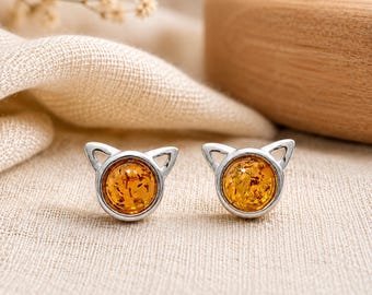 May include: A pair of silver stud earrings shaped like cat heads, each featuring a round, translucent, golden-brown amber stone set in the center. The metal frames are polished silver with pointed ears at the top and a circular base for the stone.