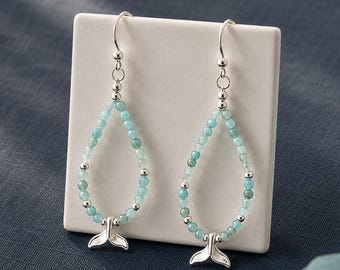 May include: A pair of dangle earrings featuring a teardrop-shaped loop of small, light blue and green gemstone beads accented with silver-toned metal beads. Each earring terminates in a silver-toned metal whale tail charm suspended from a hook.