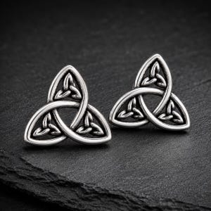 May include: A pair of silver-colored stud earrings featuring a Celtic knot design. Each earring has a three-part knot, with a central knot and three curved sections. The earrings are displayed on a dark, textured surface.