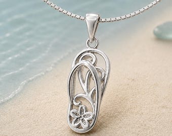 May include: A silver-toned pendant shaped like a flip-flop sandal, featuring an openwork design with swirling lines and a small five-petaled flower at the base. The pendant hangs from a delicate silver-toned box chain necklace.