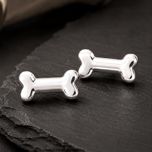 May include: Two shiny, silver-colored bone-shaped cufflinks. The cufflinks are displayed on a dark, textured surface. The design is simple and elegant, perfect for adding a touch of fun to any outfit.