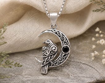 May include: A silver-toned pendant necklace featuring a crescent moon shape with intricate Celtic knotwork patterns. A stylized bird is perched on the moon, which is accented by a single small, round, black gemstone. The pendant hangs from a box chain.