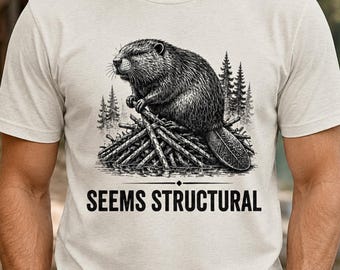May include: A cream-colored t-shirt featuring a black and white graphic of a beaver perched on a pile of logs in front of a forest. Below the illustration, the text SEEMS STRUCTURAL is printed in a bold, black, sans-serif capital font.
