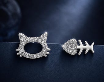 May include: A pair of silver-toned earrings featuring a cat head outline on one side and a fish skeleton on the other. Both pieces are encrusted with small, clear, sparkling stones that catch the light, creating a bright and reflective appearance.