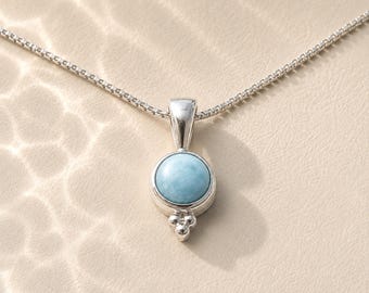 May include: A silver pendant necklace featuring a round, light blue Larimar gemstone set in a polished silver bezel. The pendant includes a small decorative cluster of three silver spheres at the base and hangs from a delicate silver box chain.