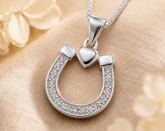 May include: A silver-toned necklace featuring a horseshoe-shaped pendant adorned with small, clear, sparkling stones. A small, smooth, silver-toned heart sits at the top center of the horseshoe, connecting it to a delicate, thin box-link chain.
