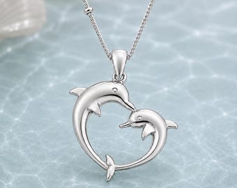 May include: A silver-toned necklace featuring a pendant shaped like two dolphins forming a heart outline. The pendant is suspended from a delicate silver-toned chain with small, evenly spaced metal beads along the length of the chain links.
