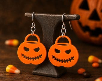 May include: A pair of bright orange dangle earrings shaped like jack-o'-lantern candy buckets. Each earring features a cutout face with triangular eyes and a jagged mouth, suspended from silver-toned metal hooks by small circular jump rings.