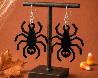 May include: A pair of dangling earrings featuring black spider-shaped charms with eight legs and two circular eyes. The spiders are suspended from silver-toned metal fishhook wires with small circular jump rings connecting the charms to the hooks.